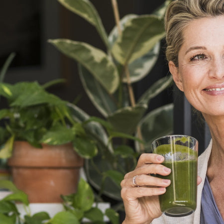 Mittelalte Frau sitzt mit grünem Smoothie in der Hand an der Terrassentür mit grünen Topfpflanzen im Hintergrund.
